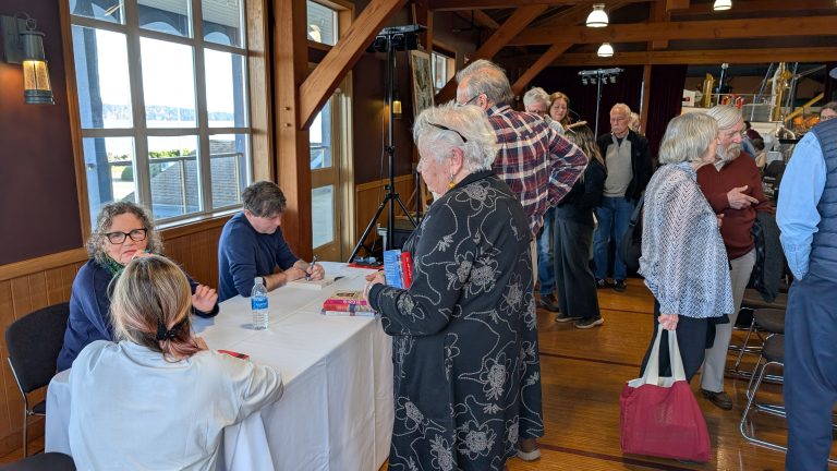 People stand in line at a book signing event. Two authors sit at a table signing books while attendees chat and wait. The scene is inside a wooden room with large windows and natural light.