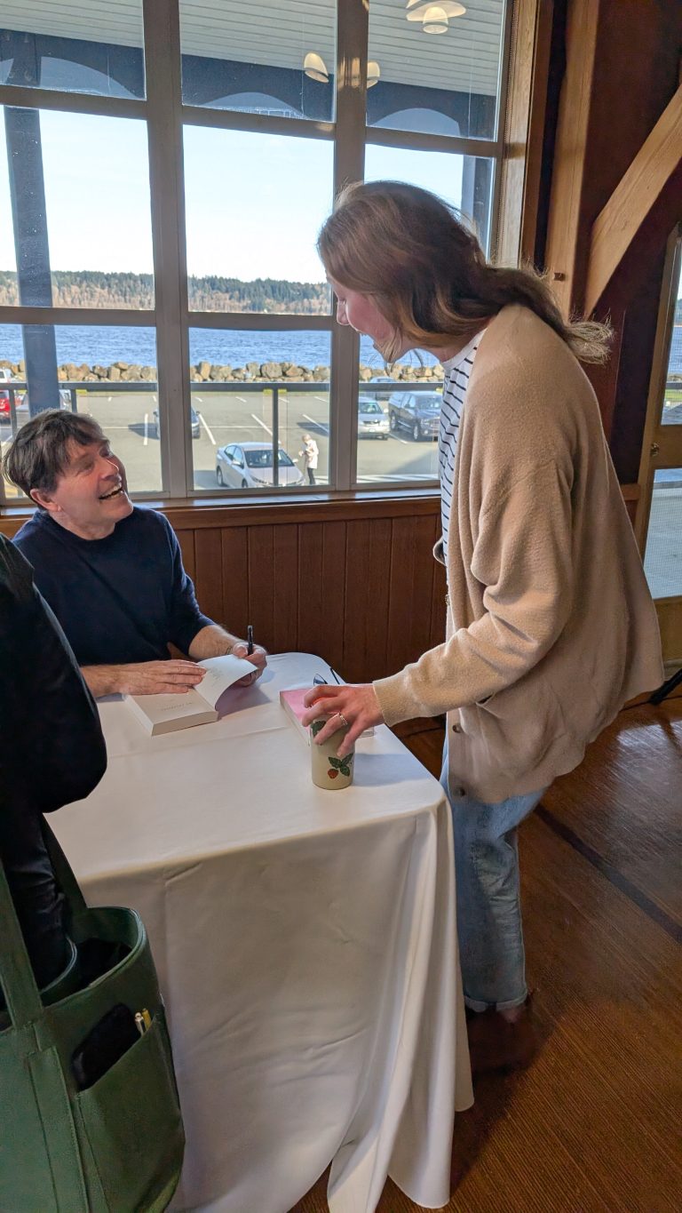 A smiling man sits at a table signing a book for a standing woman holding a coffee cup. Large windows behind them show a scenic view of water, trees, and blue sky.