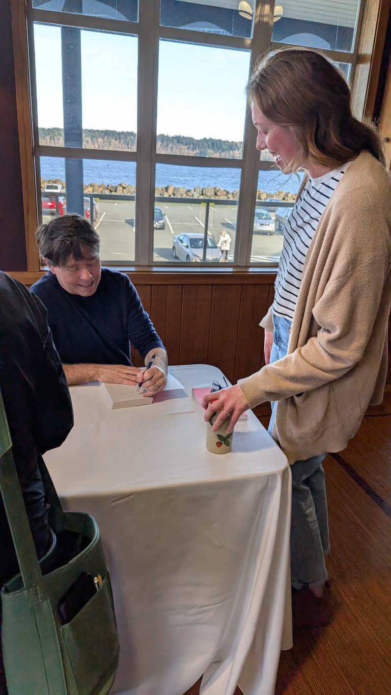A man sits at a table signing a book for a woman who stands holding a cup. Large windows behind them show a scenic outdoor view with water, trees, and parked cars.