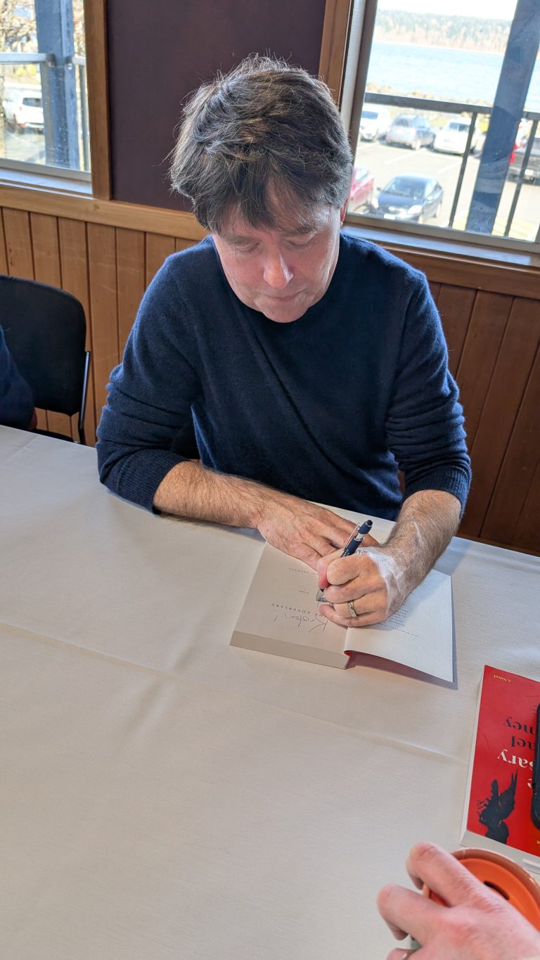 A person with dark hair is seated at a table, signing a book. A red book is visible on the table, and another hand is holding a cup in the lower right corner. Large windows show cars and trees outside.