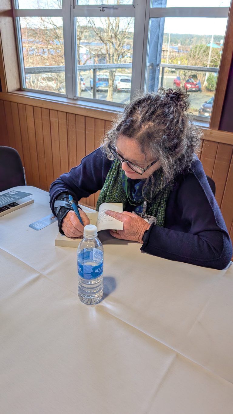 A person with curly hair and glasses is sitting at a table, signing a book. A water bottle and laptop are on the table. Large windows show parked cars and trees outside.