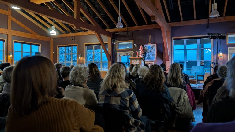 A woman stands at a podium speaking to a seated audience in a rustic, wood-beamed room with large windows at dusk.