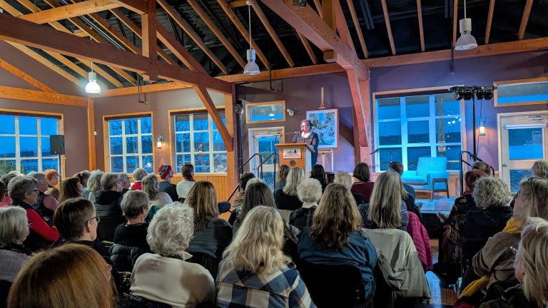 A speaker addresses an attentive audience in a wooden hall with exposed beams and large windows at dusk. The room is filled with seated people facing the podium.