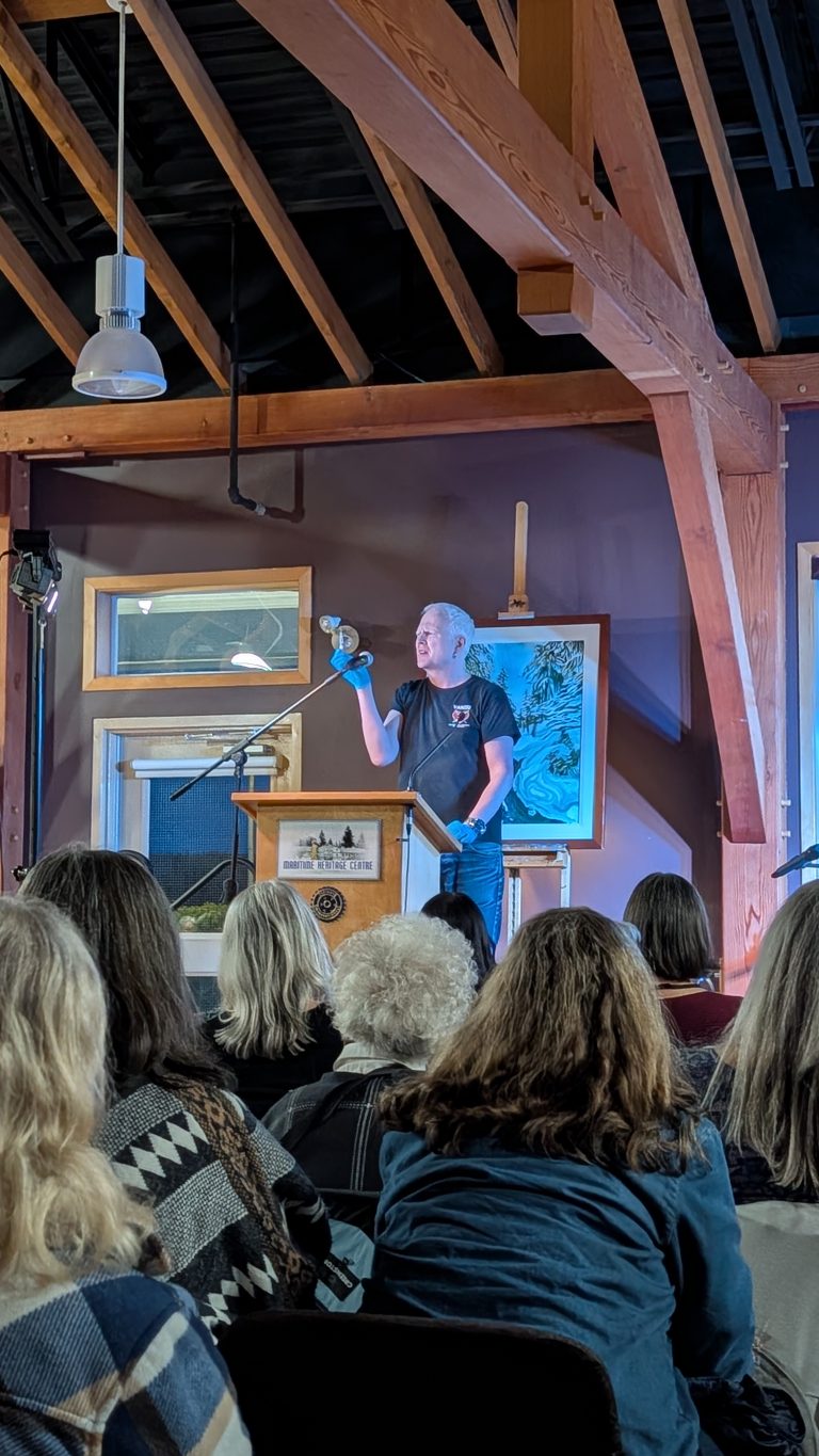 A person stands on stage holding up a bird, speaking to an audience seated indoors. Wooden beams, art, and windows are visible in the background.